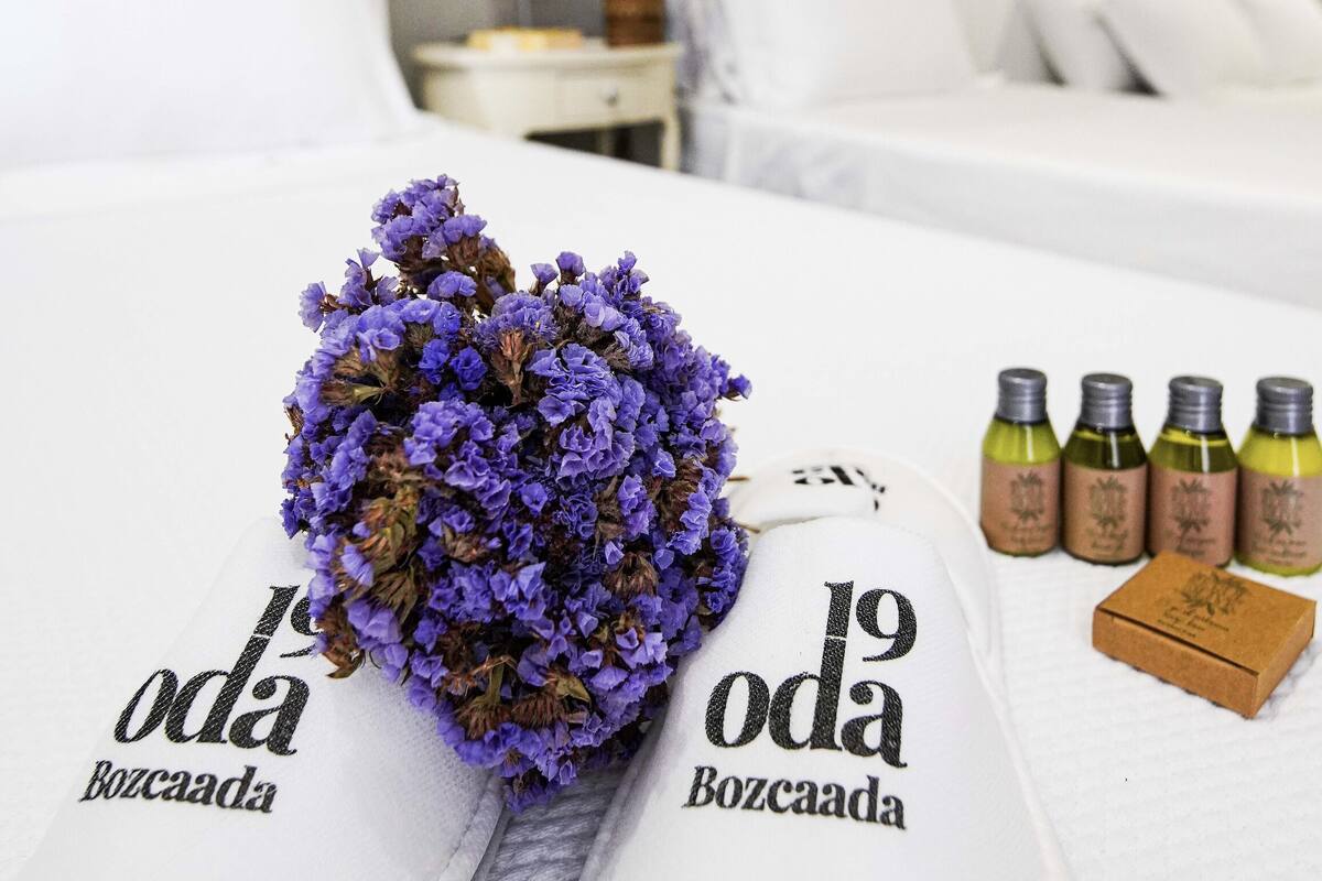 A bed with a bouquet of purple flowers, a white pillow, and four bottles of wine on a white table.