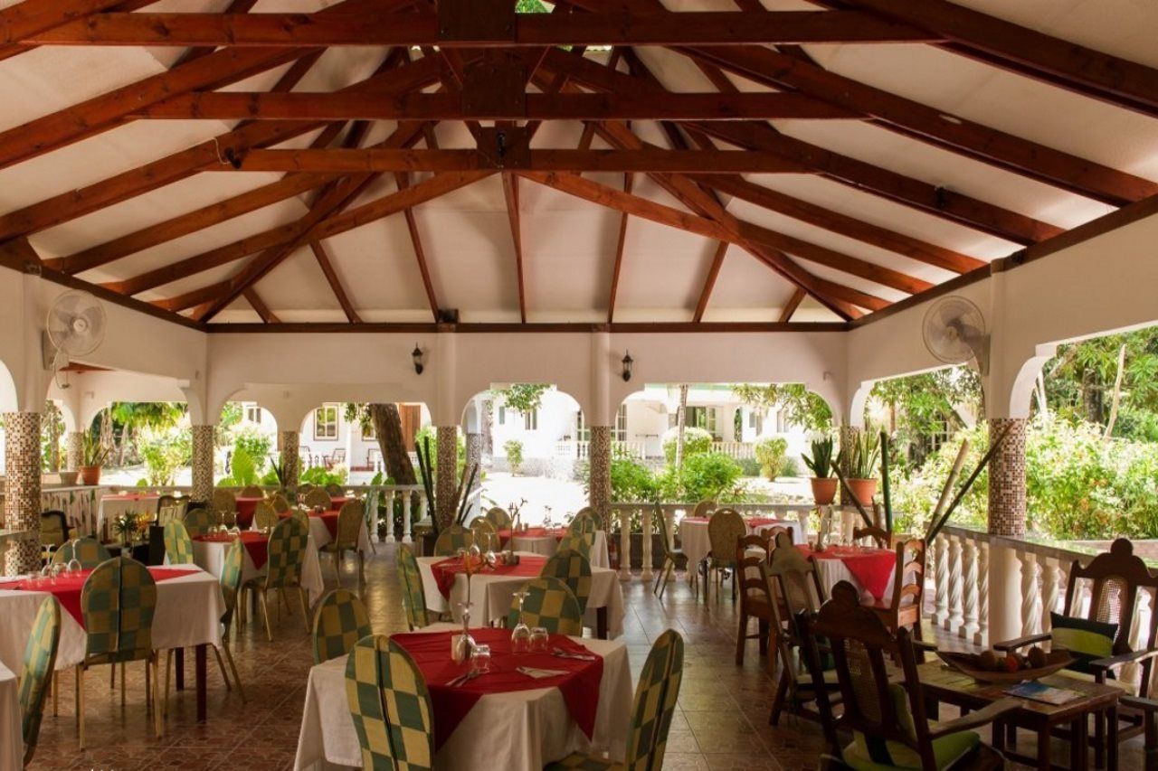 An indoor dining area with tables and chairs, a high ceiling with wooden beams, and a view of greenery outside.