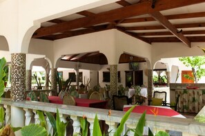 An outdoor dining area with tables and chairs, covered by a wooden roof structure, and surrounded by columns and arches.