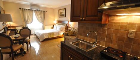 A hotel room with a bed, a desk, a chair, a dining table, a kitchen area with a sink and stove, and a window with curtains.