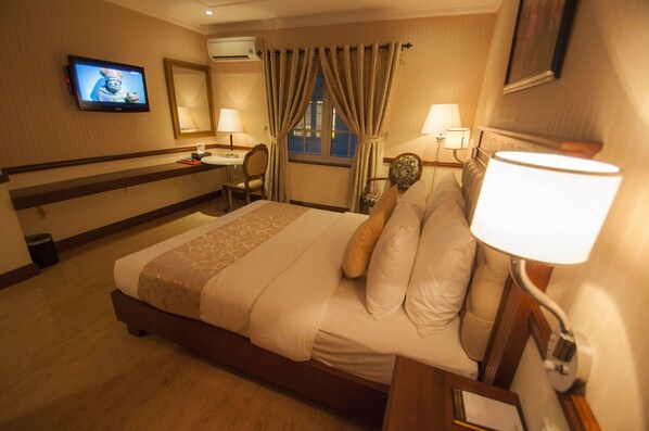 A hotel room with a bed, bedside table, lamp, desk, chair, TV, and curtains.