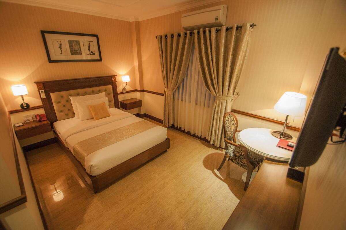 Hotellihuone, jossa on suuri sänky, kaksi yöpöytää valaisimineen, pyöreä pöytä ja tuoli sekä peili.