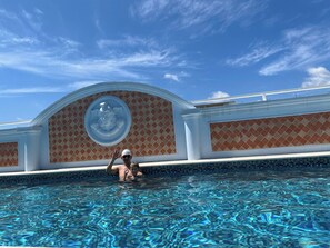 Une piscine avec une eau d’un bleu limpide, une personne dans la piscine et, en arrière-plan, un bâtiment dont la façade présente un motif décoratif.