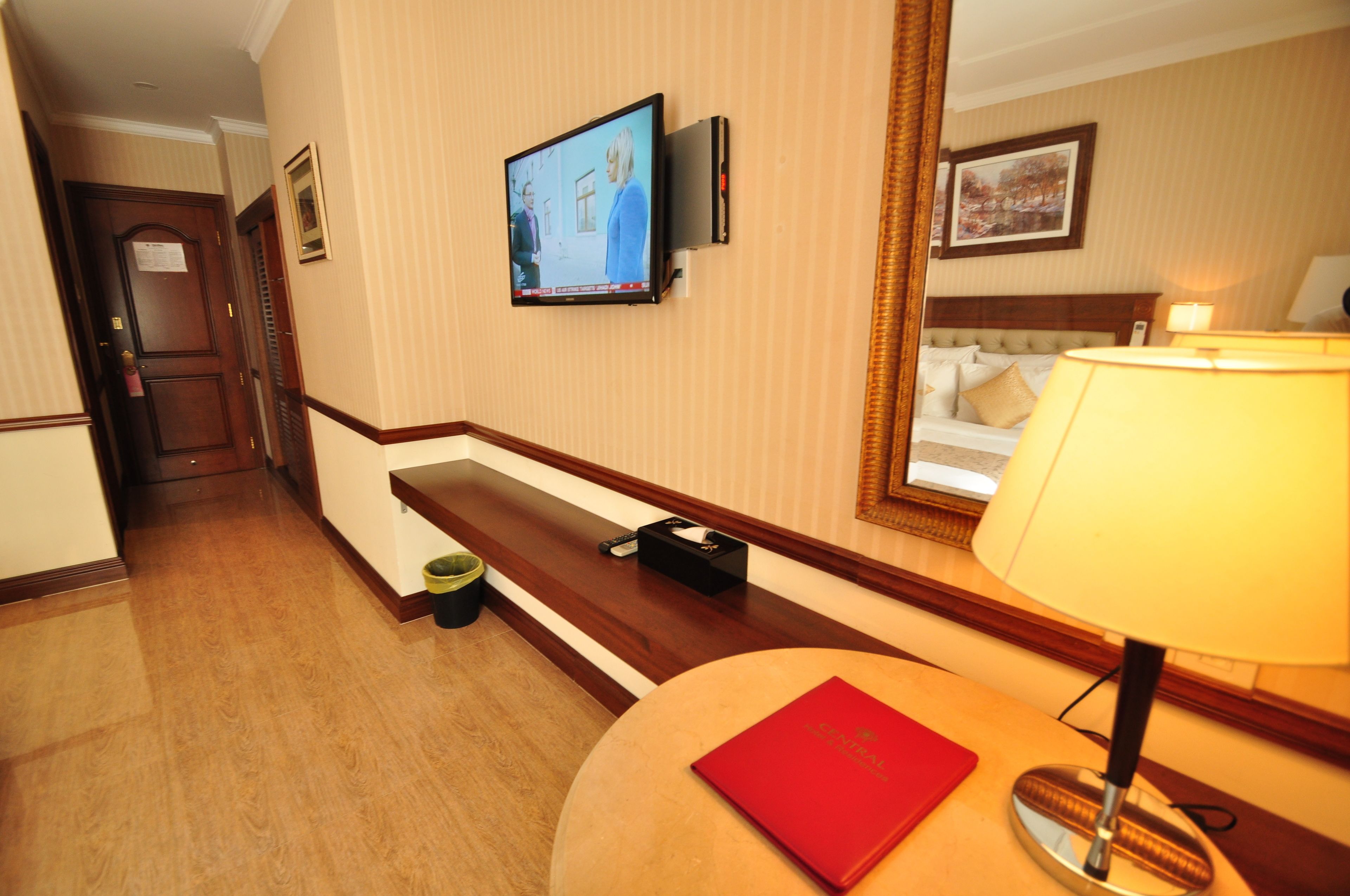 Ein Hotelkorridor mit an der Wand montiertem Fernseher, einer Bank, einem Nachttisch mit Lampe und einem Bett mit Kopfteil.