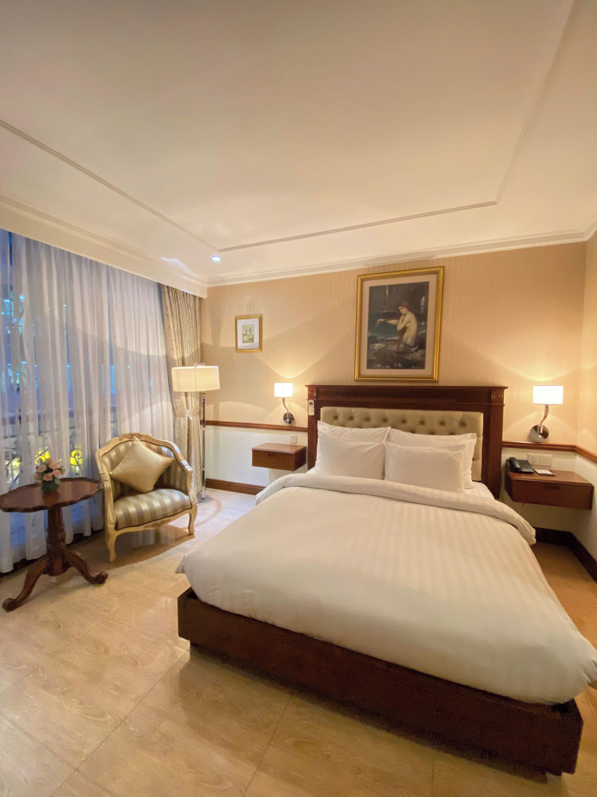 A hotel room with a large bed, a chair, a bedside table, a nightstand, and a lamp.