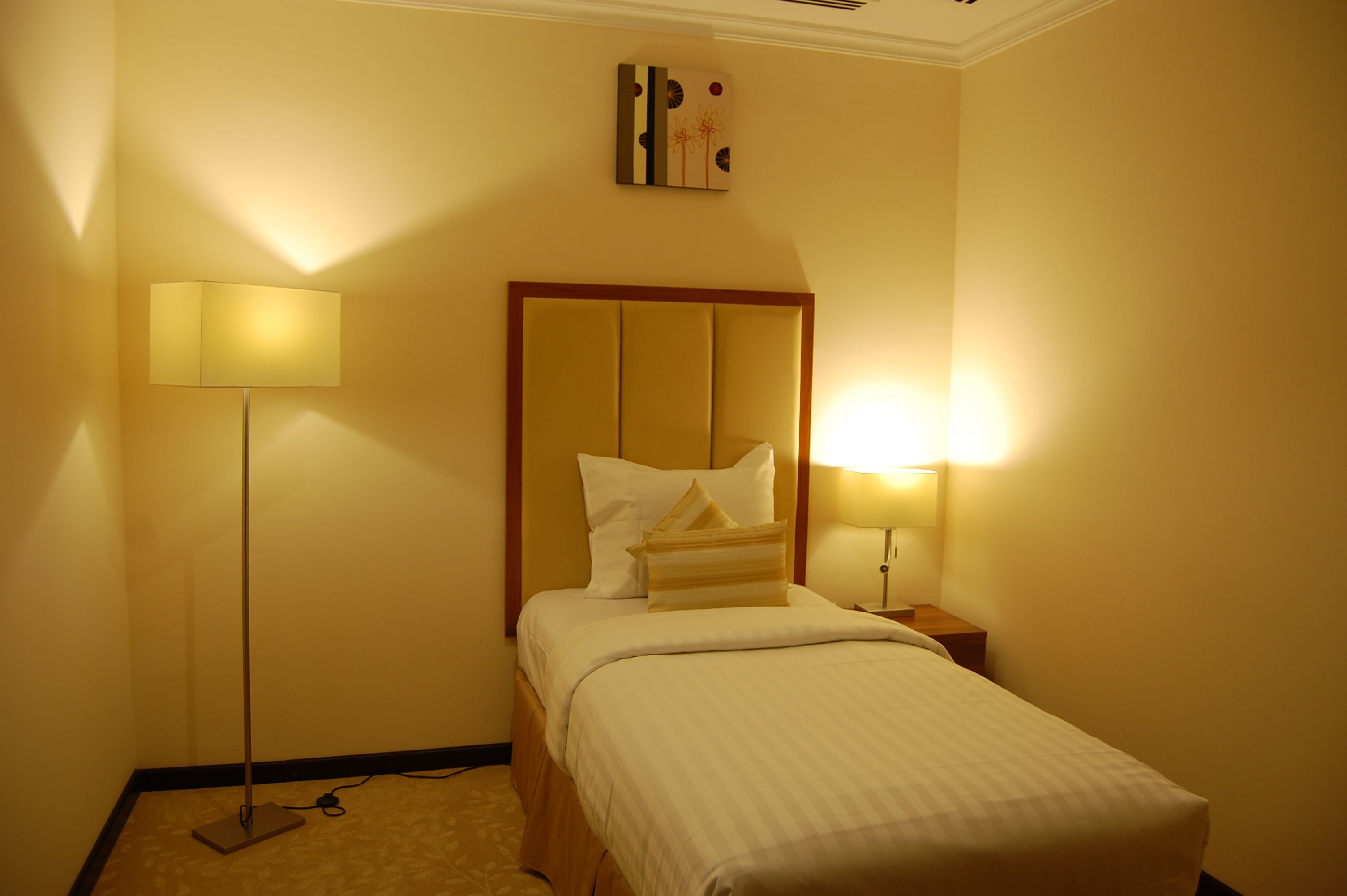 Una habitación de hotel con cama, una lámpara de noche, una lámpara de pie y una obra de arte colgada en la pared.