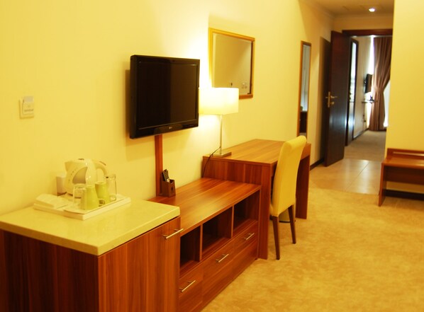 A hotel room with a wooden desk, a television mounted on the wall, a mirror, and a chair.