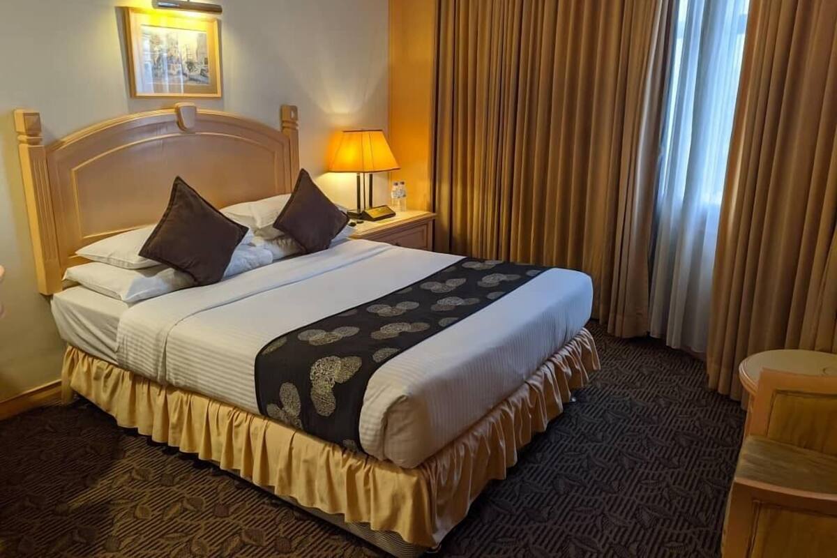 Une chambre d’hôtel avec un grand lit, une table de chevet avec une lampe, un tableau encadré au mur et une fenêtre avec des rideaux.