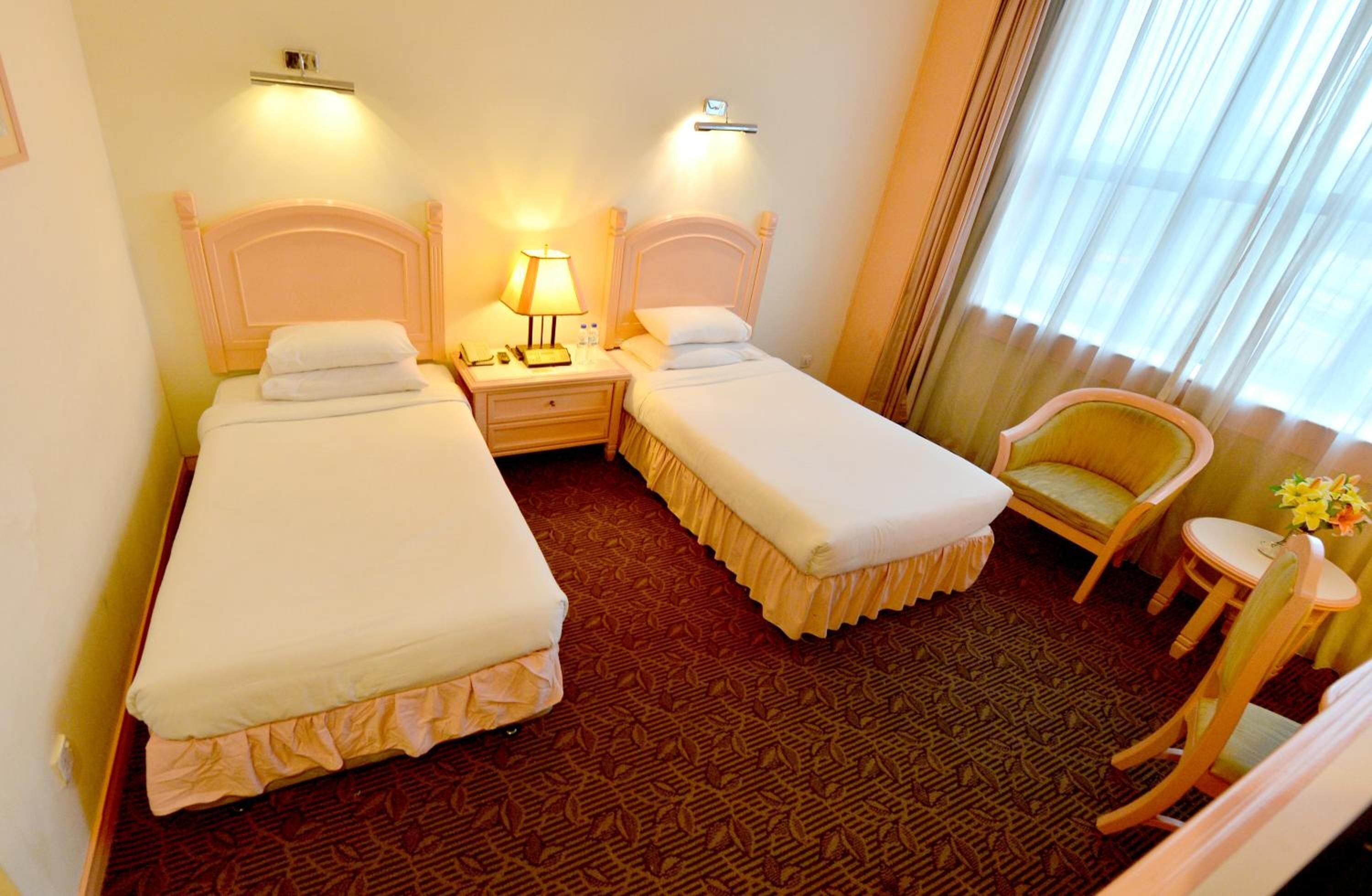 A hotel room with two single beds, a nightstand with a lamp, a chair, a small table with a vase, and a window with curtains.