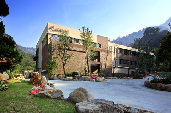 A modern building with a stone facade, large windows, and a spacious entrance area with a concrete pathway and rock landscaping. - Goya Spring Resort (Taichung)