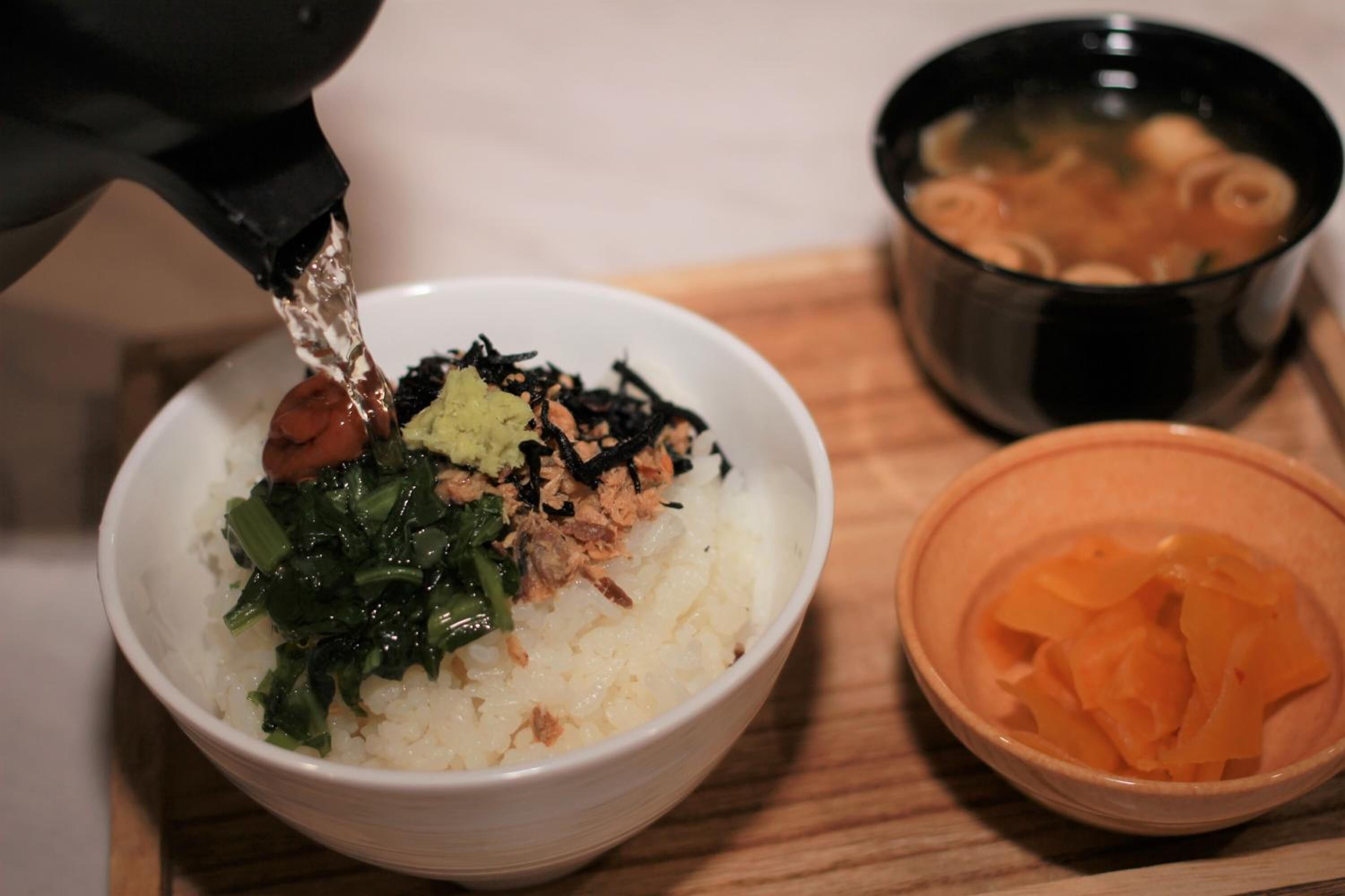 A bowl of rice with toppings, a bowl of soup, and a bowl of pickled vegetables on a wooden table.