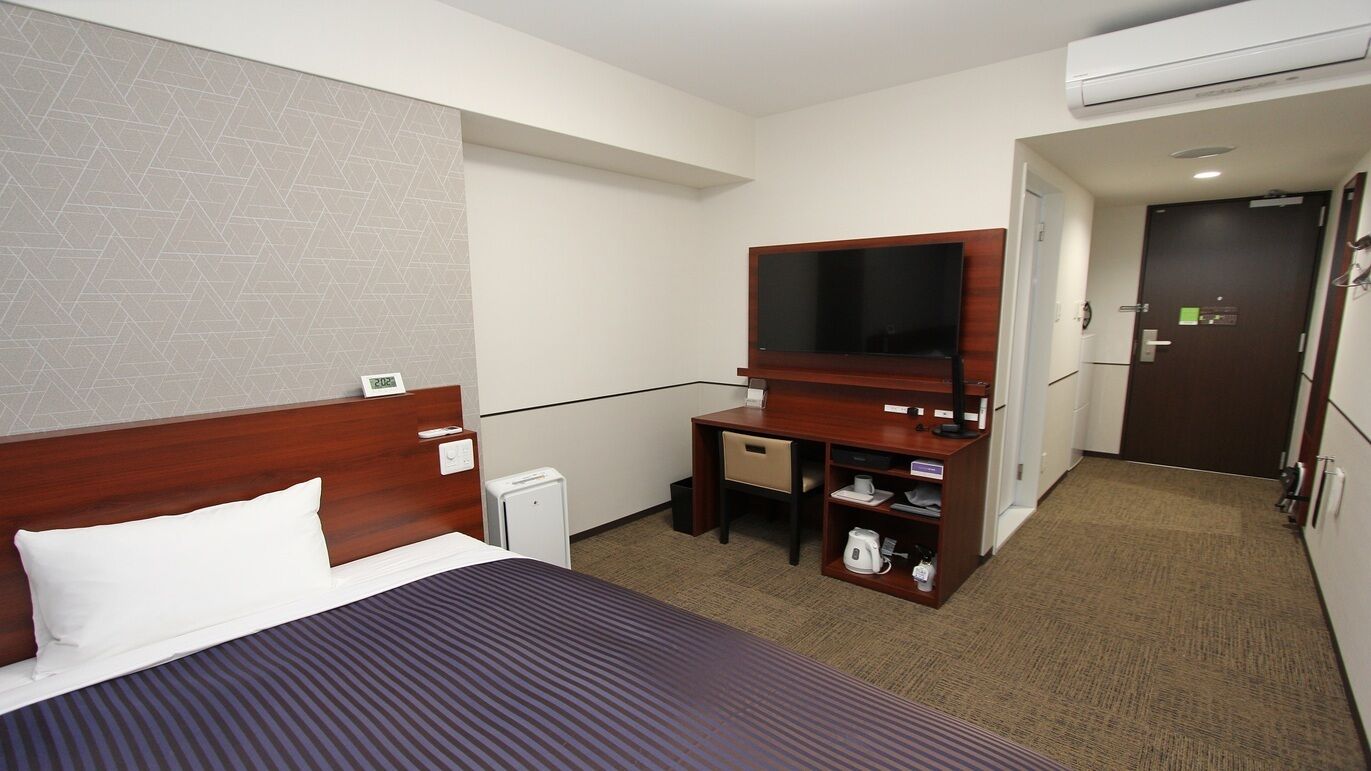 Een hotelkamer met een bed, een bureau, een televisie, een kleine koelkast en een deur naar een andere kamer.