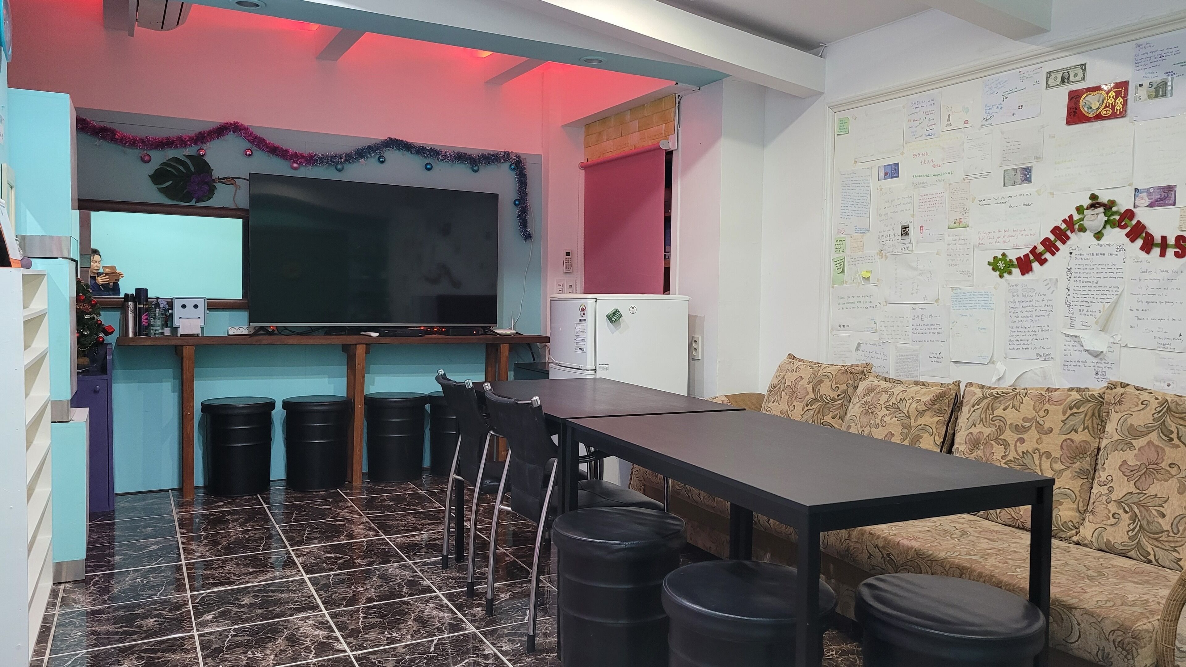 Una habitación con una mesa larga, sillas, un sofá, un televisor, un refrigerador y una pared decorada con notas.