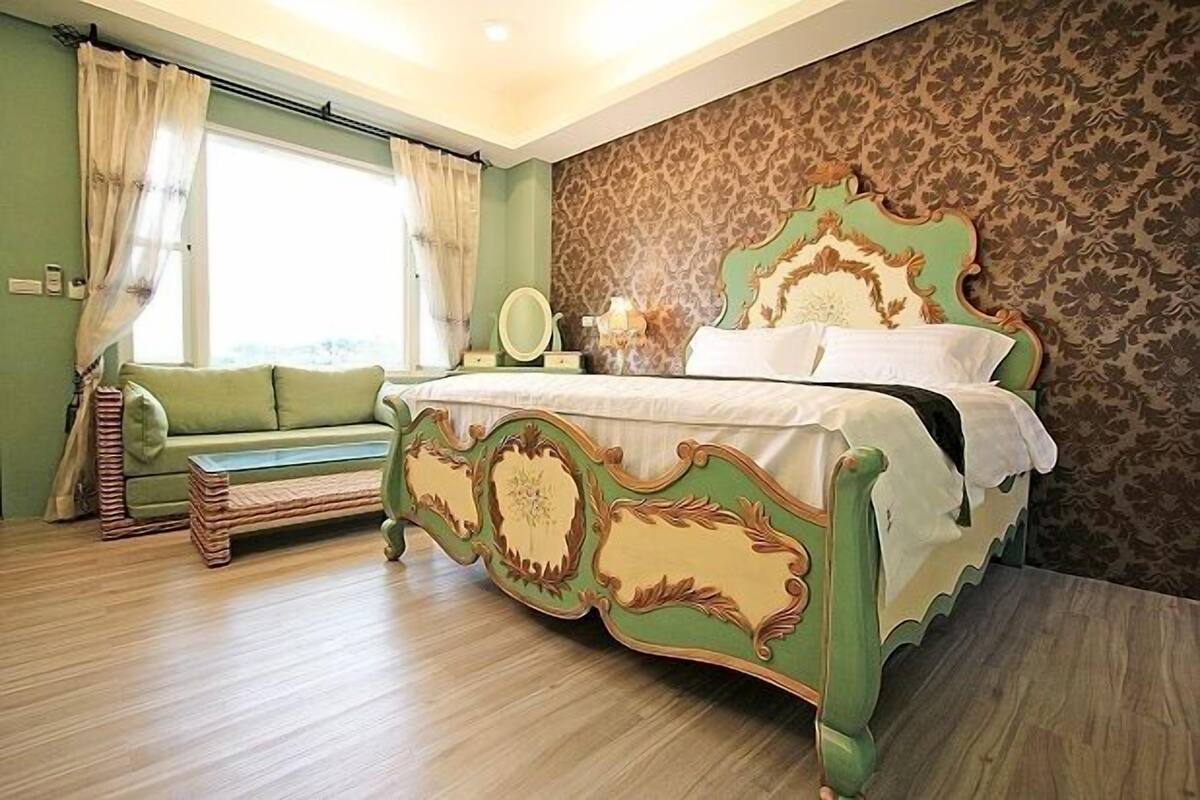Quarto com sofá estofado verde, piso de madeira, papel de parede estampado e cama com cabeceira verde.
