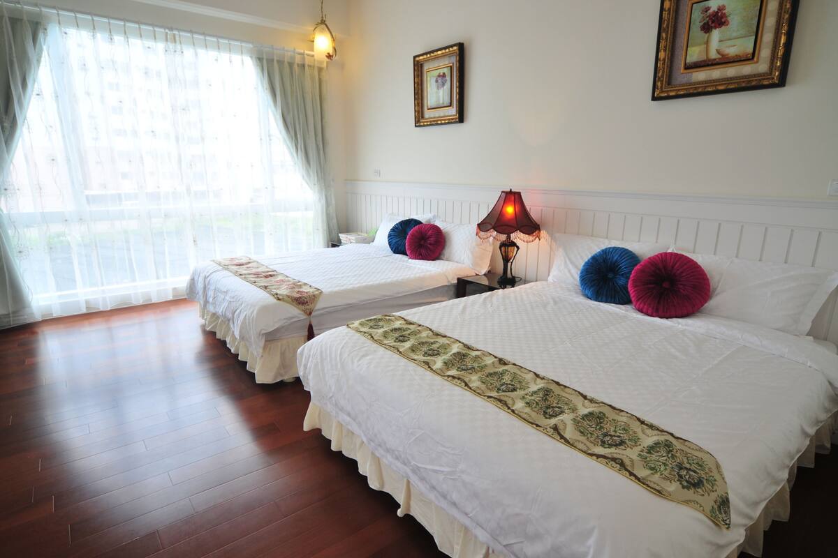 Habitación de hotel con dos camas, una ventana con cortinas translúcidas, una lámpara de noche y un cuadro enmarcado en la pared.