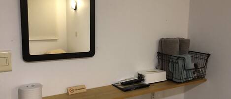 A bathroom with a mirror, a shelf with a 'No Smoking' sign, a basket, a toilet paper roll, a remote control, and a kettle.