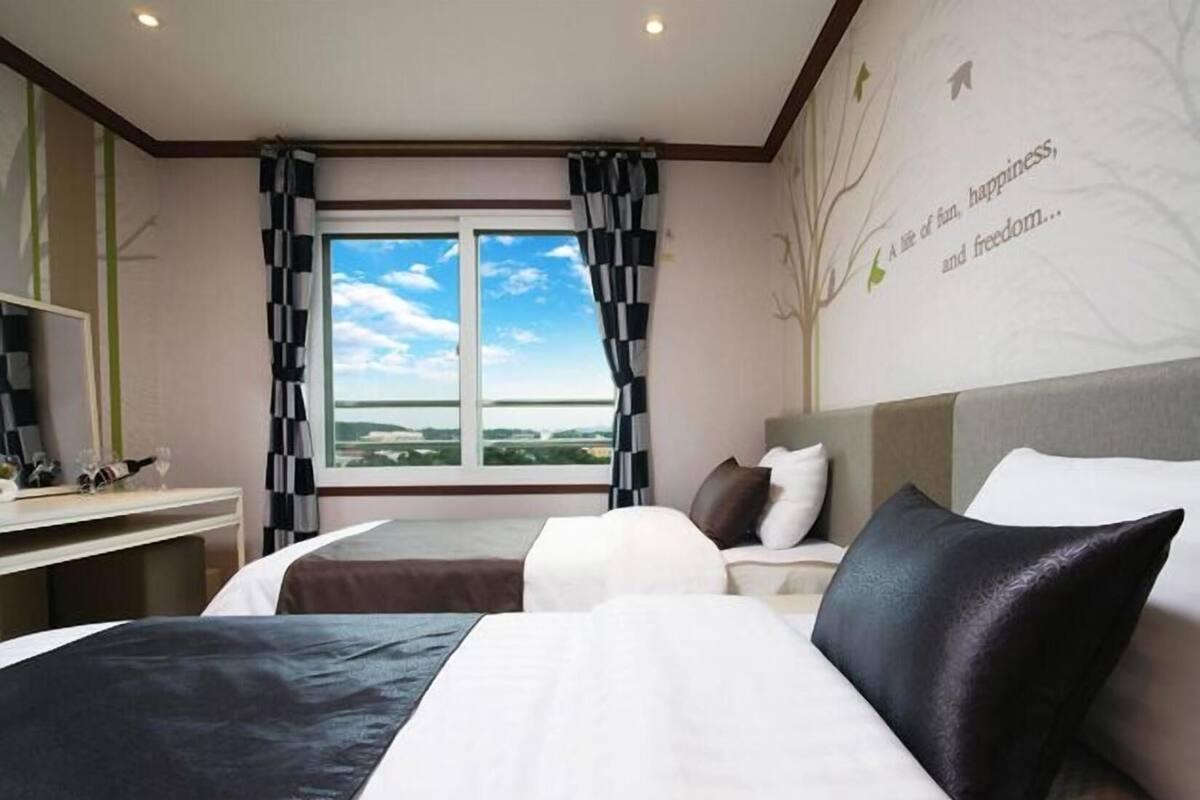 Habitación de hotel con dos camas, un escritorio con una silla, un ventanal con cortinas, y una pared con un árbol y texto.