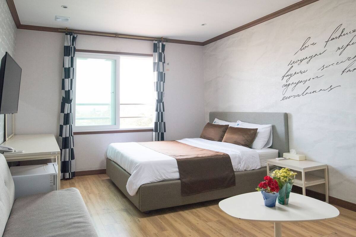 Habitación de hotel con cama, mesita de noche, una pequeña mesa redonda con flores, televisión y ventana con cortinas.