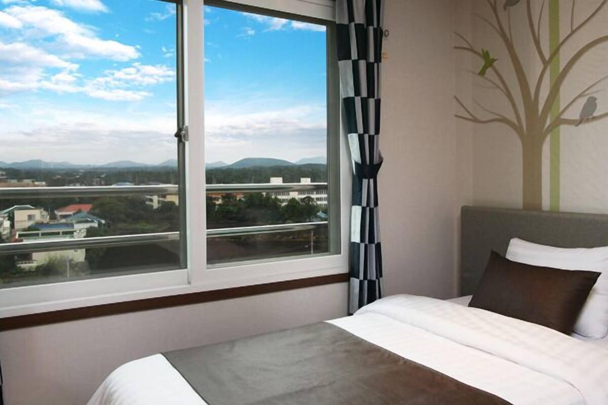Habitación de hotel con una cama, vistas de la ciudad a través de un ventanal grande y un mural en la pared con un árbol y pájaros.