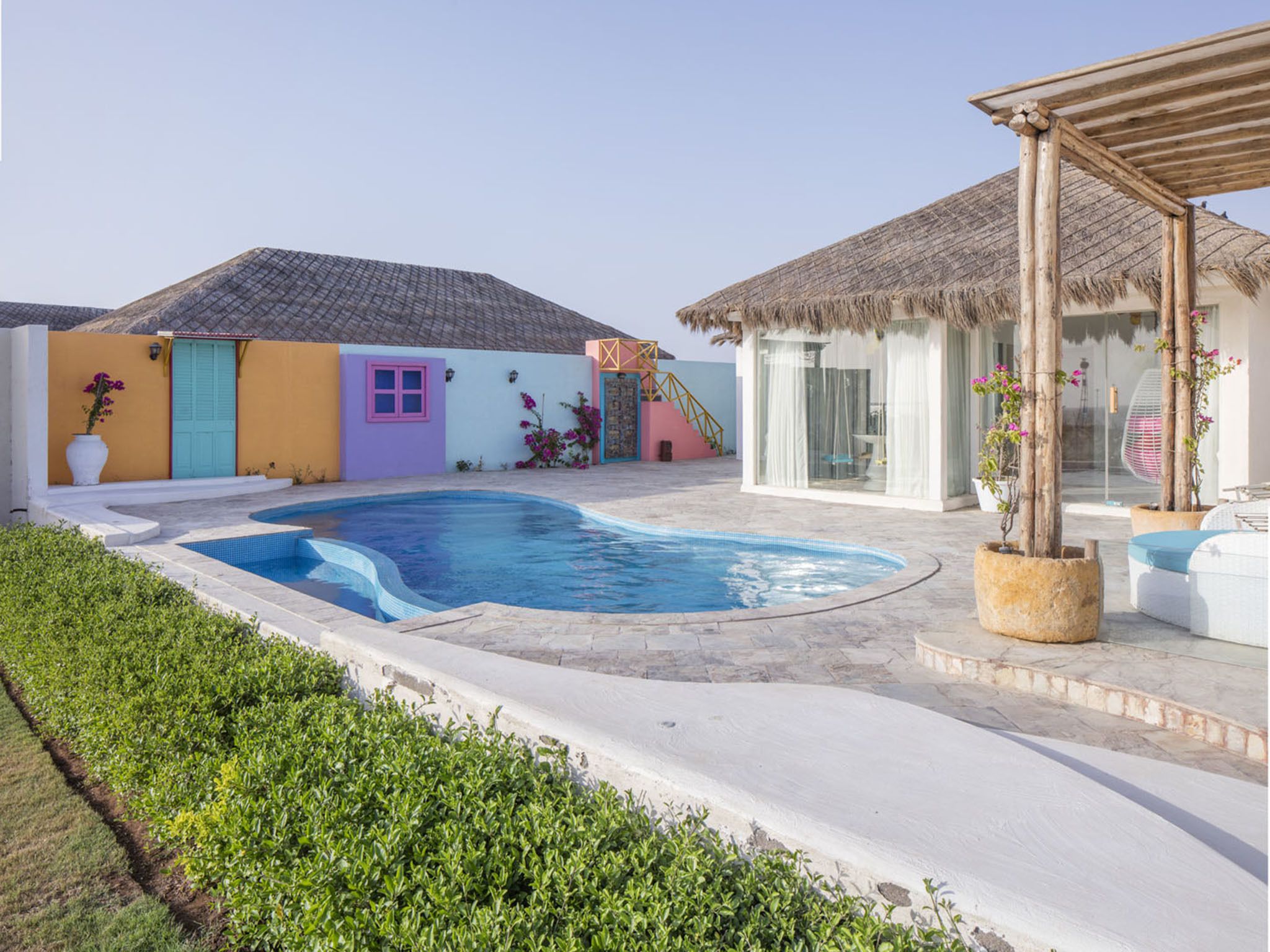 A swimming pool with a curved edge, surrounded by a white patio area. There are colorful buildings with doors and windows, and a thatched-roof structure.