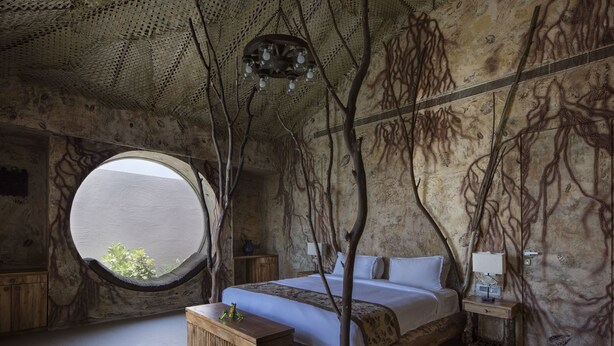A bedroom with a round window, a bed, a wooden nightstand, and a wall with tree-like patterns. - Serena Beach Resort (Mandvi)