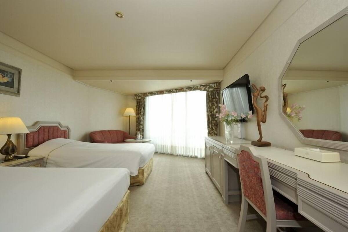 Ein Hotelzimmer mit zwei Betten, einem Schreibtisch, einem Stuhl, einem großen Spiegel und einem Fenster mit Vorhängen.