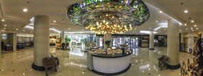Eine geräumige Lobby mit einer zentralen Rezeption, einer großen Glasdecke im Stil des Jugendstils und einem polierten Boden.