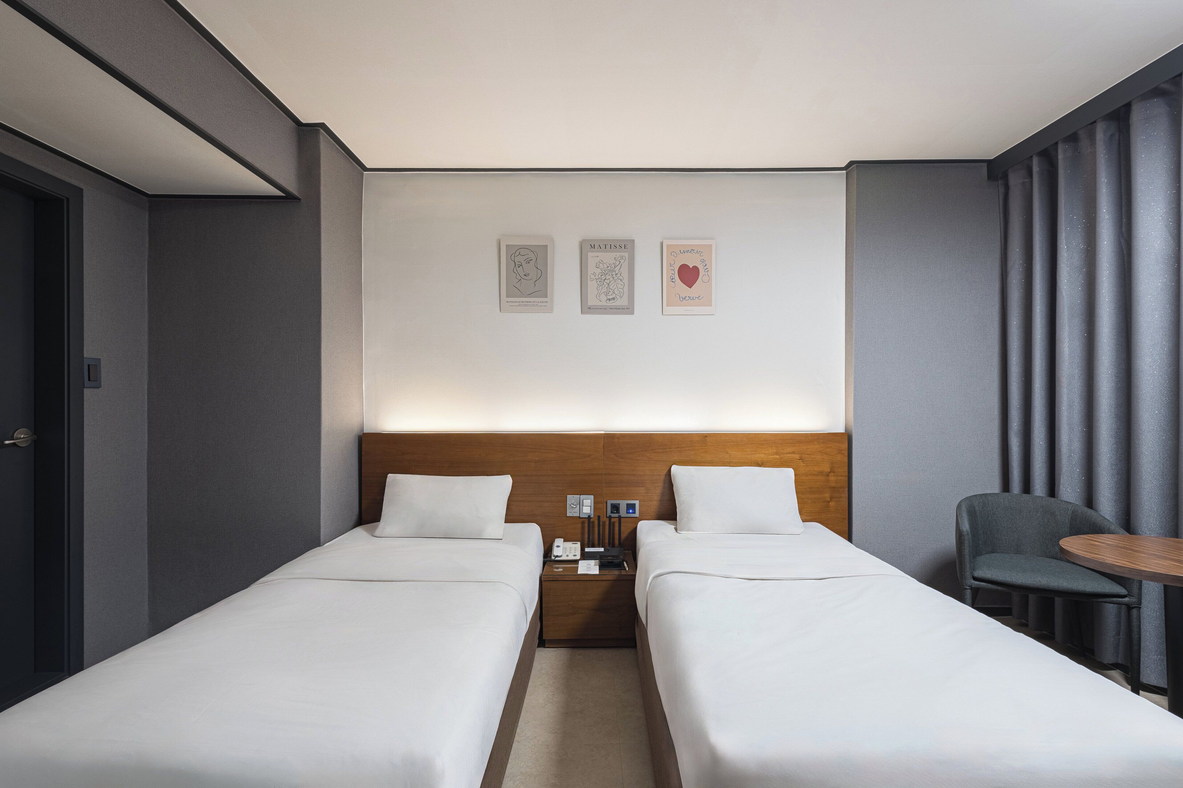 A hotel room with two single beds, a wooden headboard, a small table, and a chair.