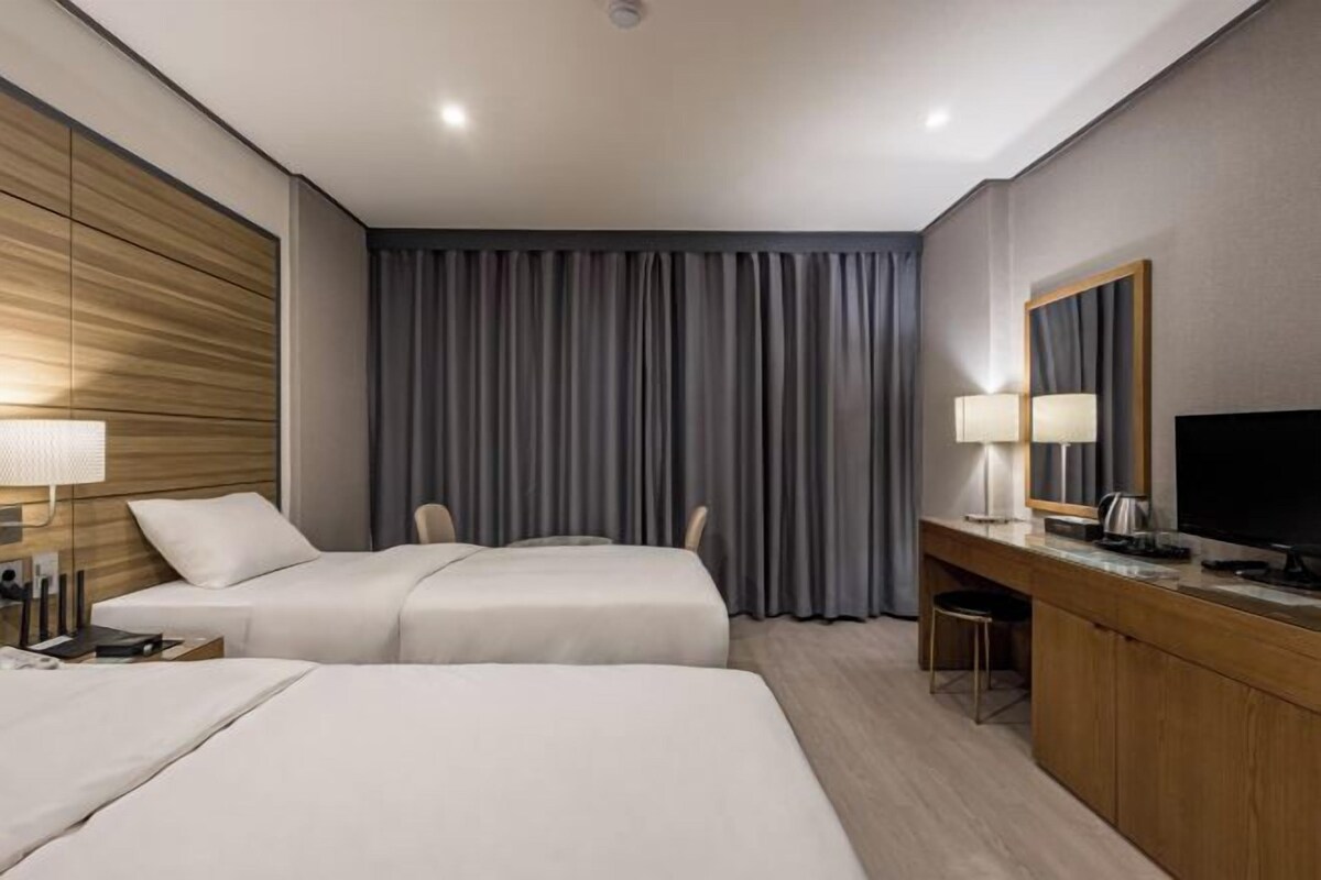 Ein Hotelzimmer mit zwei Betten, einem Schreibtisch, einem Sessel, einem Fernseher und einem großen Fenster mit Vorhängen.