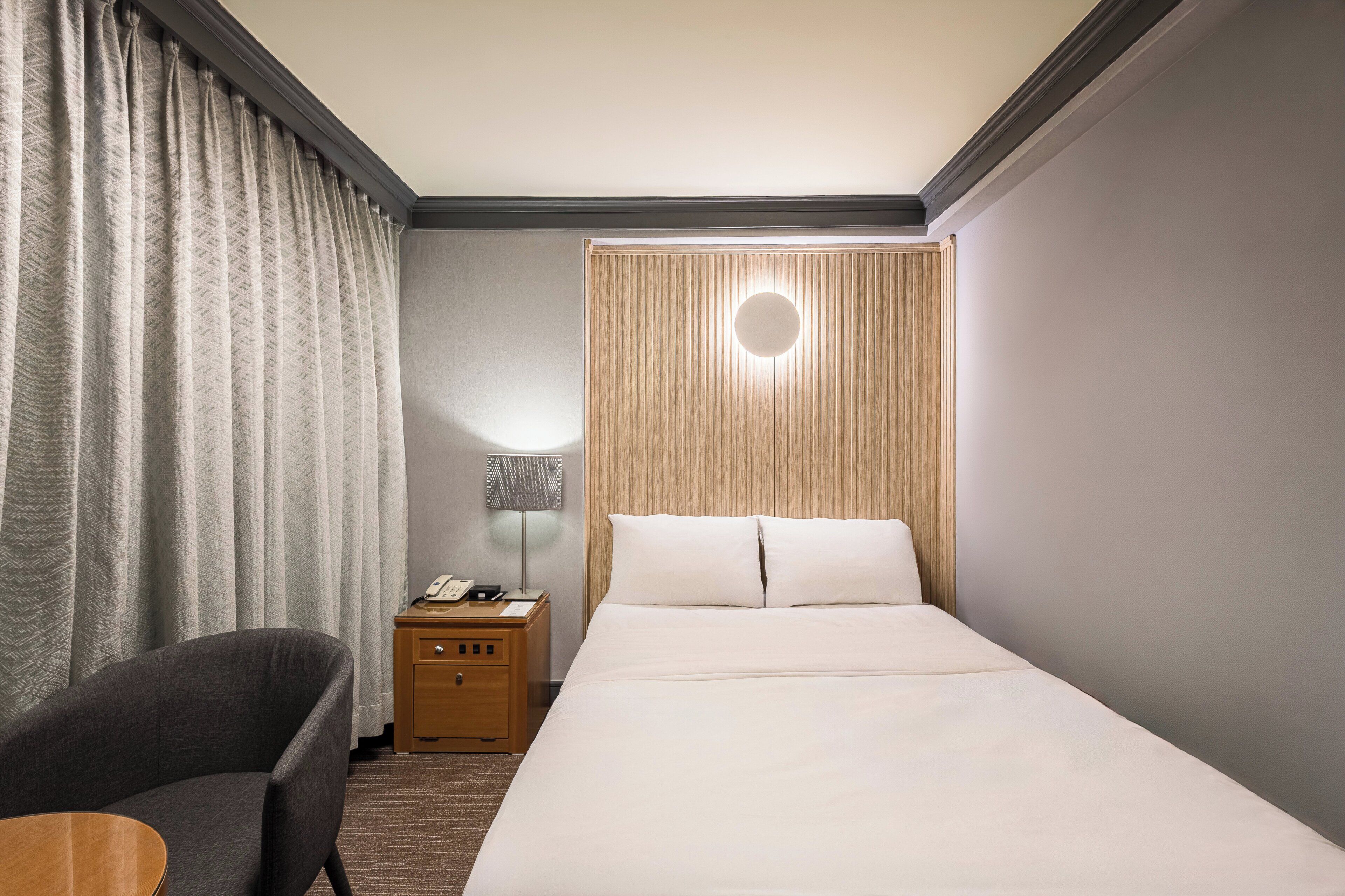 A hotel room with a bed, a chair, a nightstand, a lamp, and a window with curtains.