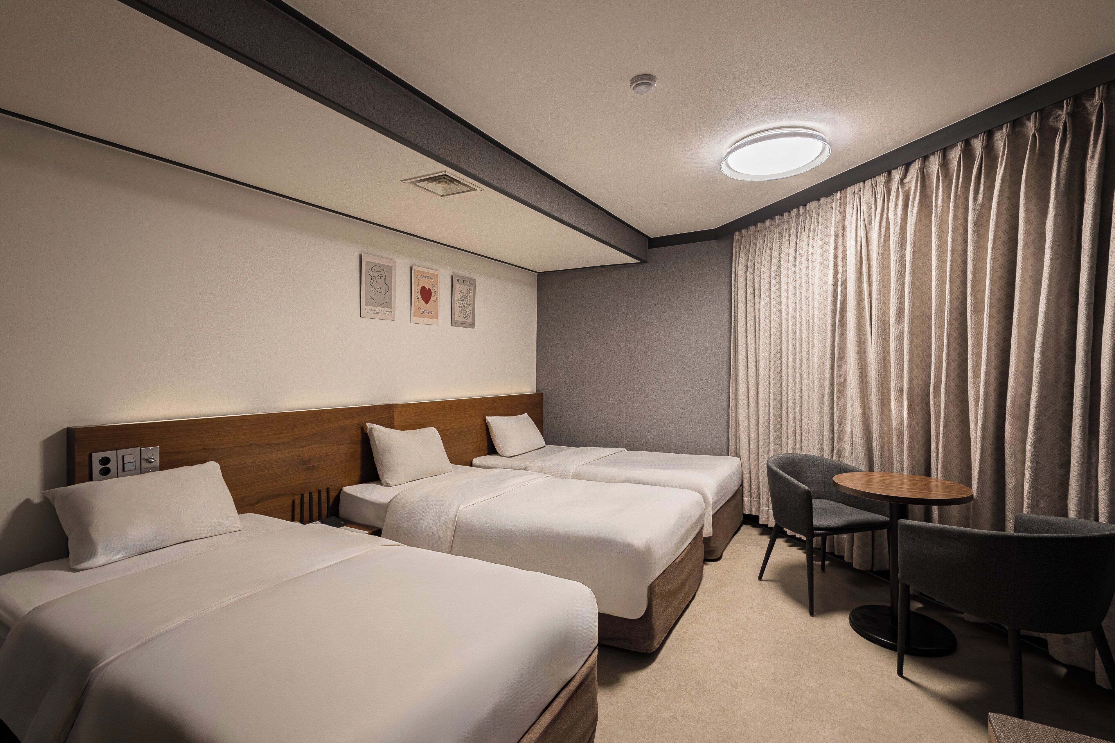 A hotel room with two beds, a small table, a chair, and a window with curtains.