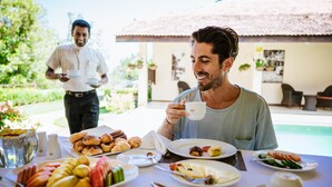A dining table with various food items and a person holding a cup, with a pool and outdoor seating in the background. - Hanthana Boutique Villa by Amaya (Kandy)