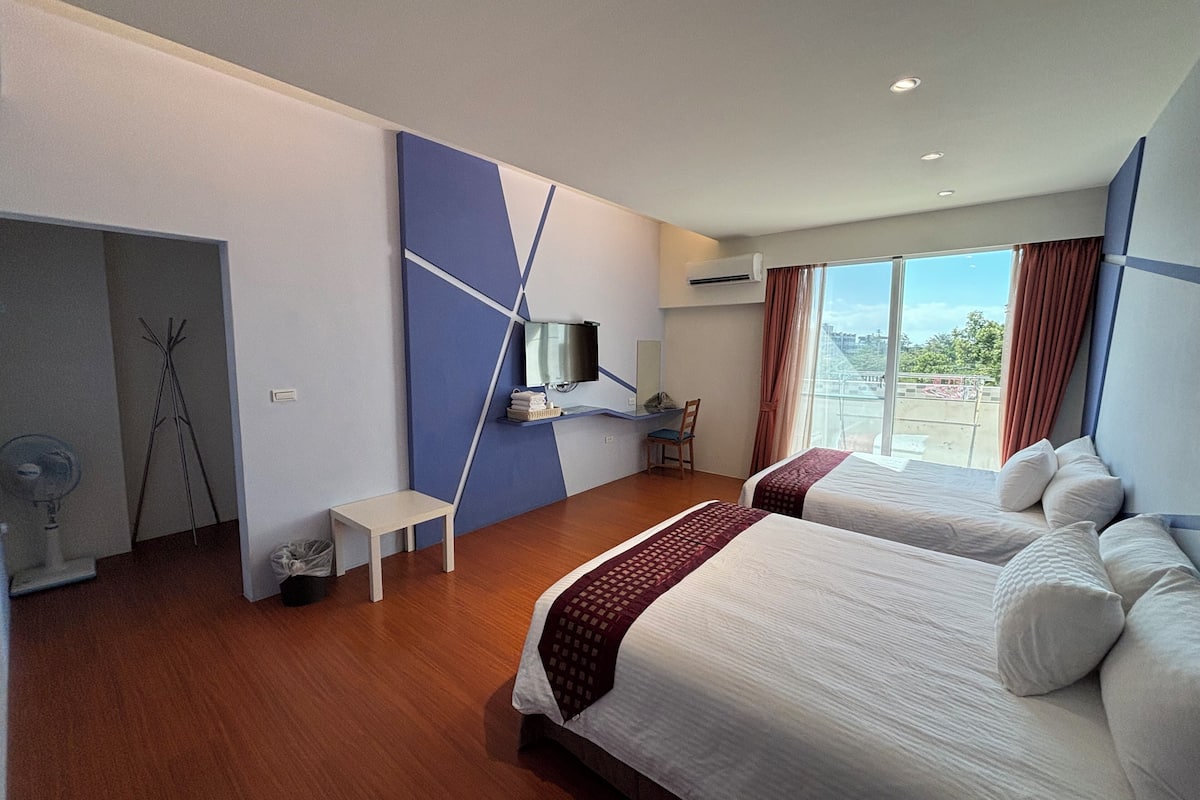 A modern hotel room with two beds, a small table, a TV, and a view of the city.