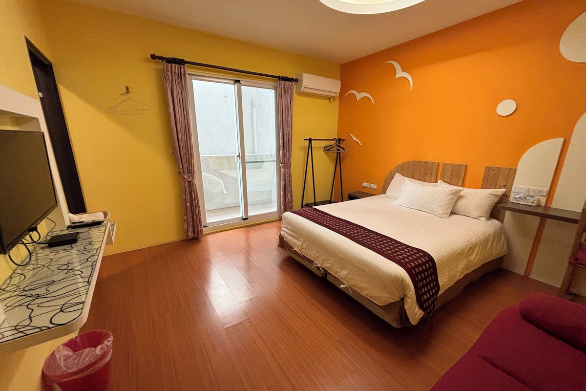 A hotel room with a bed, a desk with a computer, a TV, a window with curtains, and a wall with a bird mural.