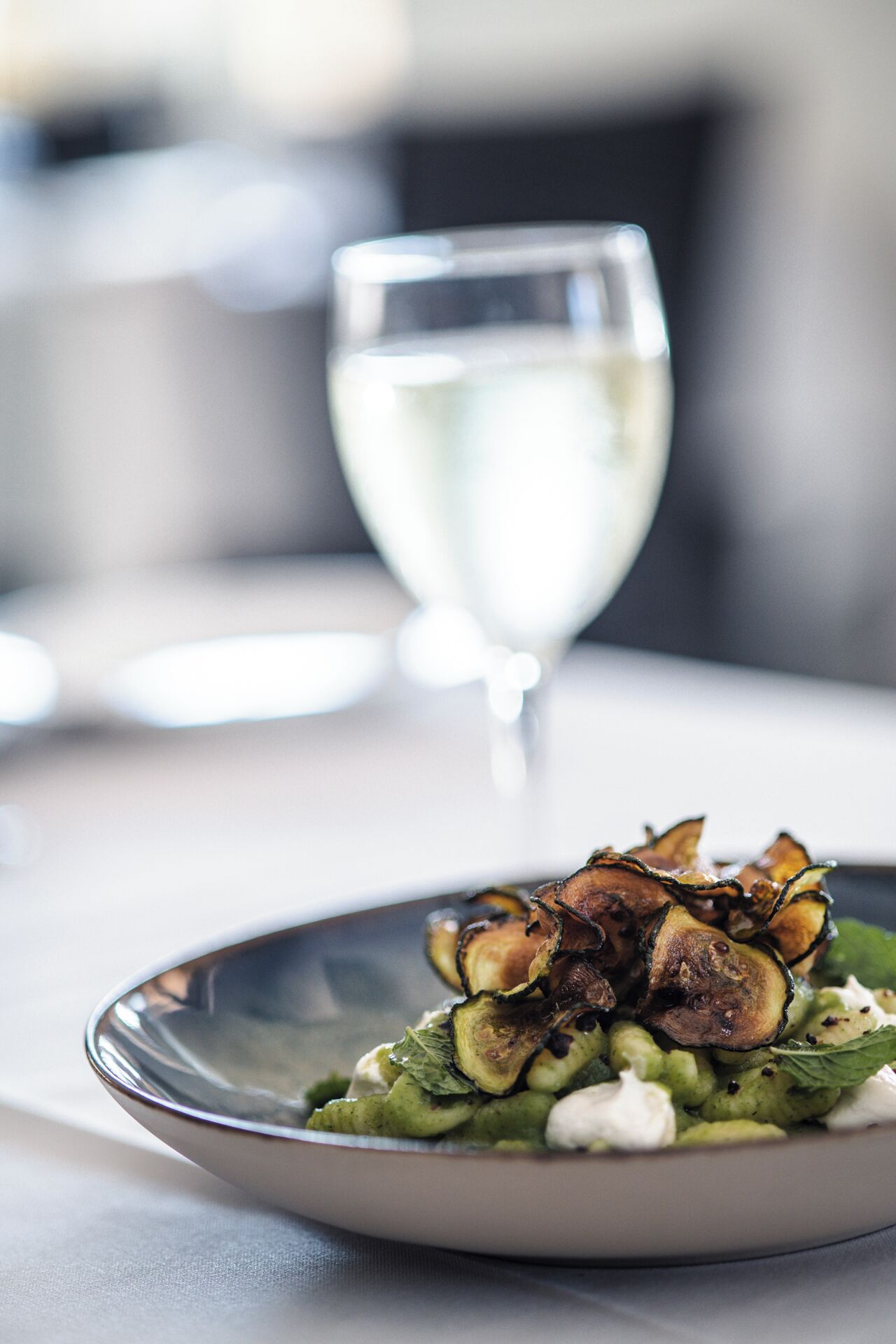 Een bord eten met geroosterde aubergine en groenten, vergezeld van een glas witte wijn op een tafel.
