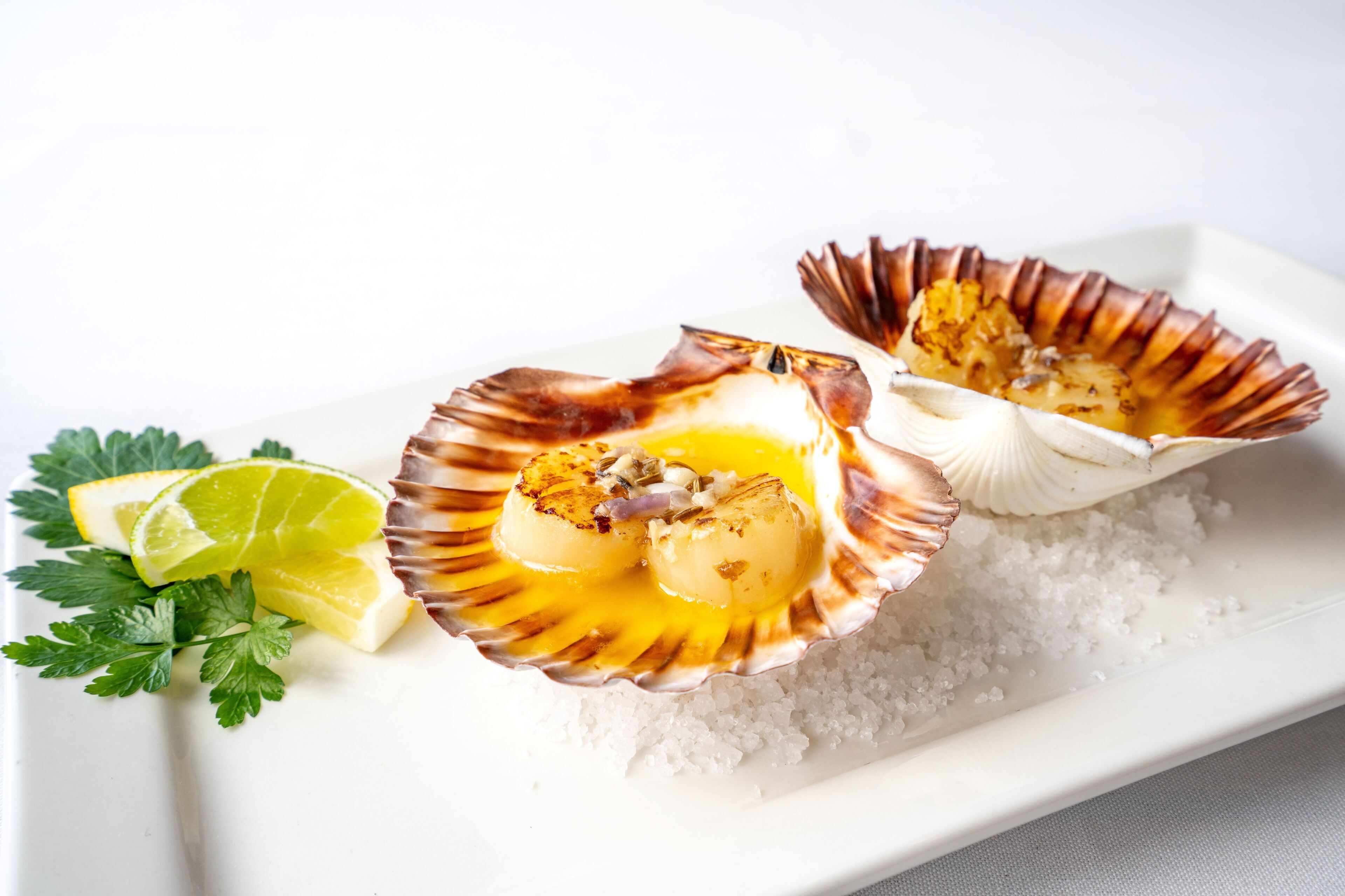 A plate with two scallops on a bed of salt, accompanied by a lime wedge and parsley.