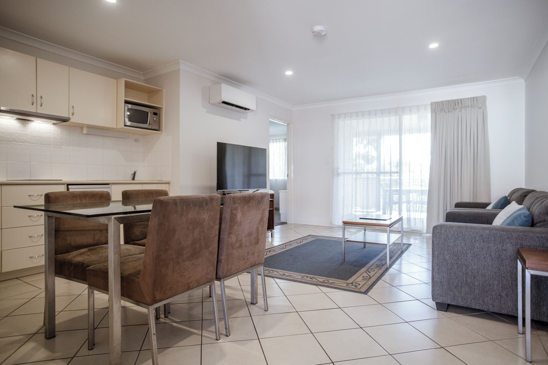 Een moderne woonkamer met een glazen eettafel, twee bruine stoelen, een grijze bank en een televisie.