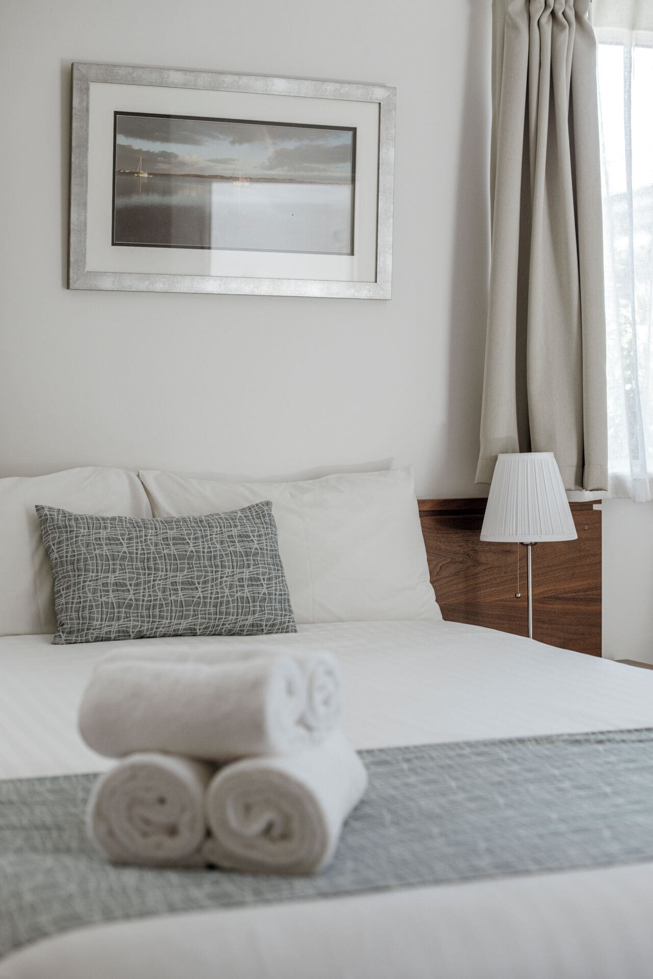 A neatly made bed with white linens and rolled towels, a framed picture on the wall, a bedside lamp, and a window with curtains.
