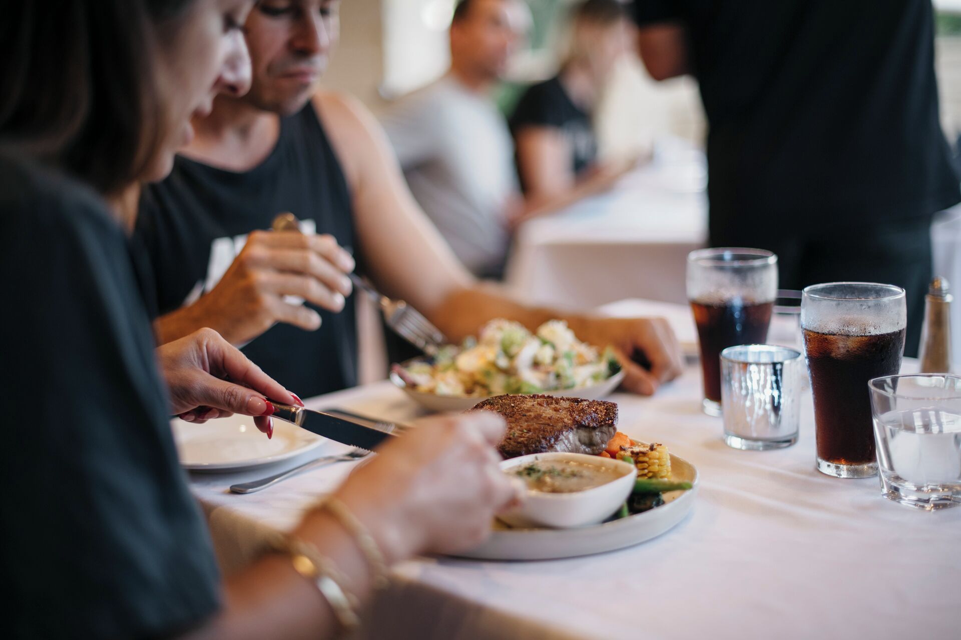 A dining table with plates of food, glasses of a dark beverage, and a person using a knife.