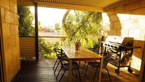 Een terras buiten met een houten tafel en stoelen, een barbecue en uitzicht op een straat en bomen.