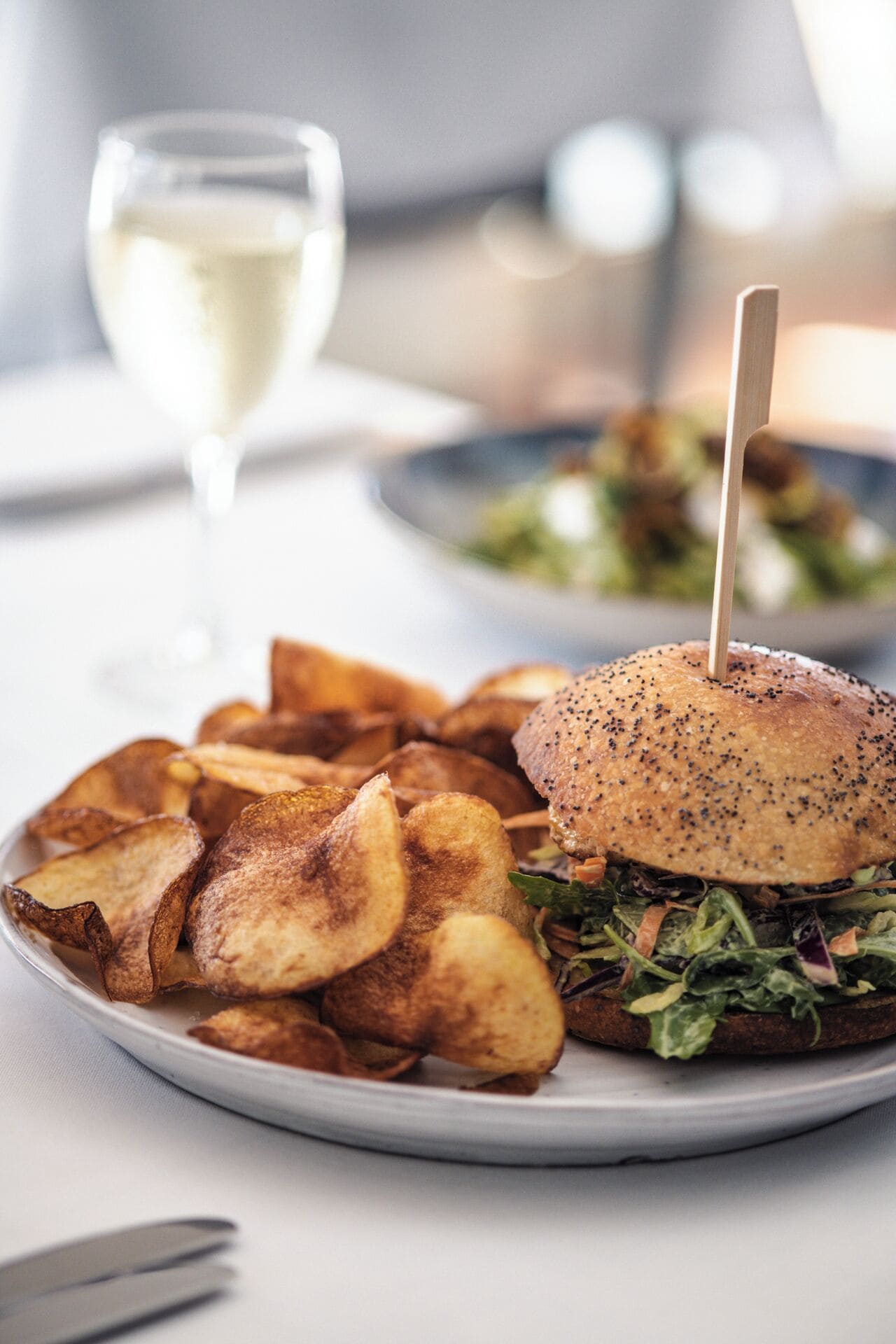A plate with a burger and potato wedges, a glass of white wine, and a blurred background.