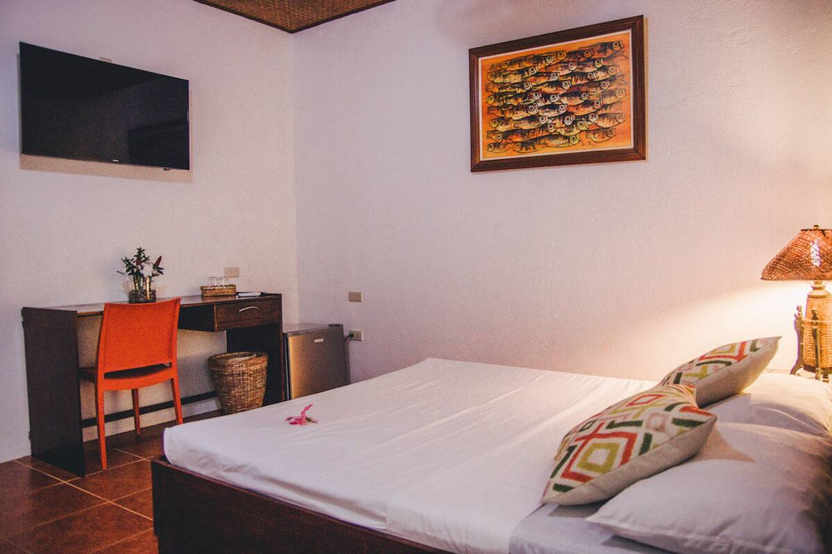 Un dormitorio con cama, escritorio, silla, televisor y una obra de arte en la pared.