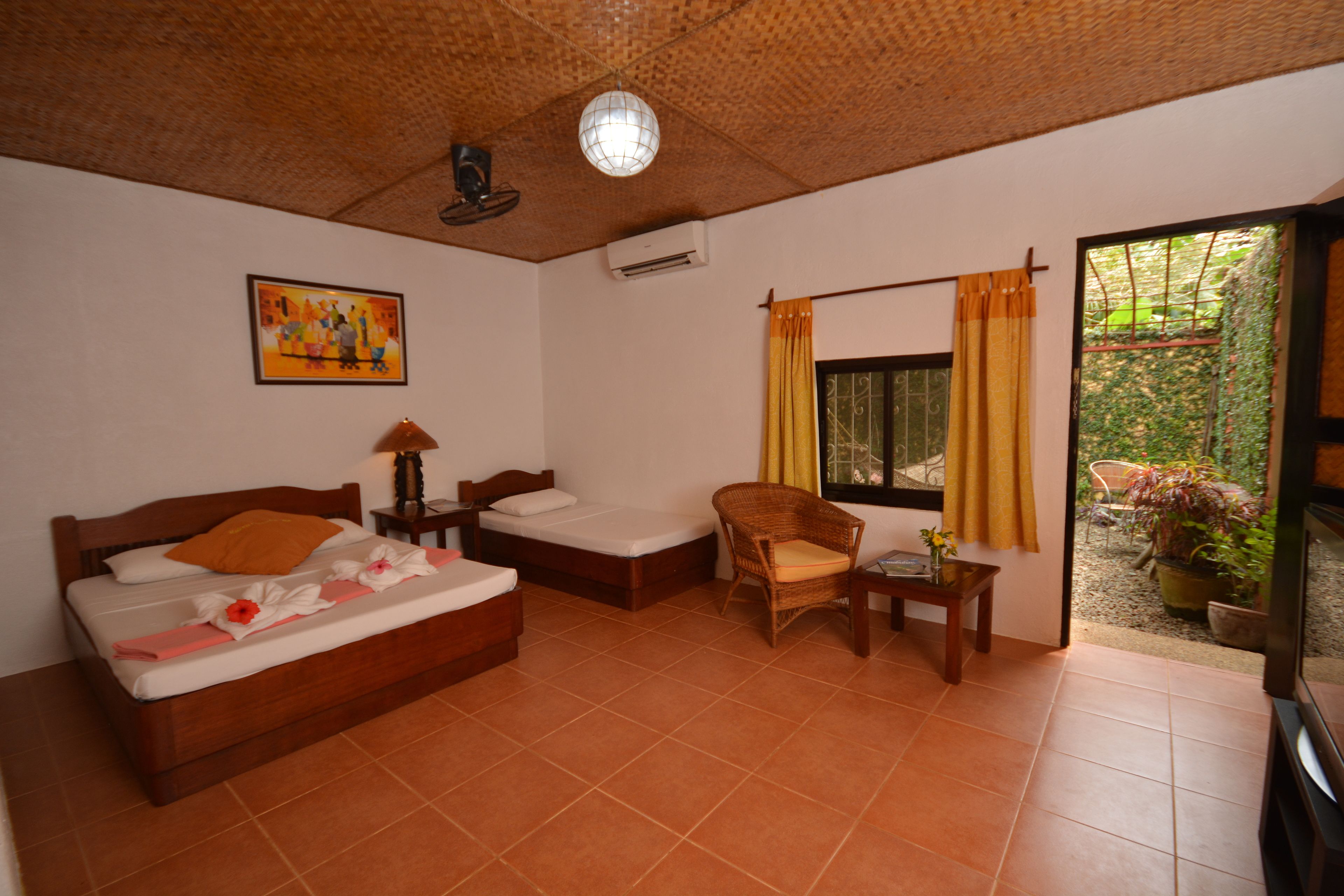 Una habitación con cama, una silla, una mesa, un ventanal con cortinas, un cuadro en la pared y un equipo de aire acondicionado.