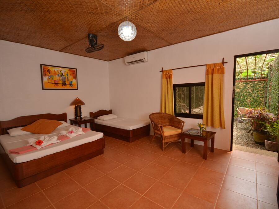 A room with a bed, a chair, a table, a window with curtains, a painting on the wall, and an air conditioning unit.