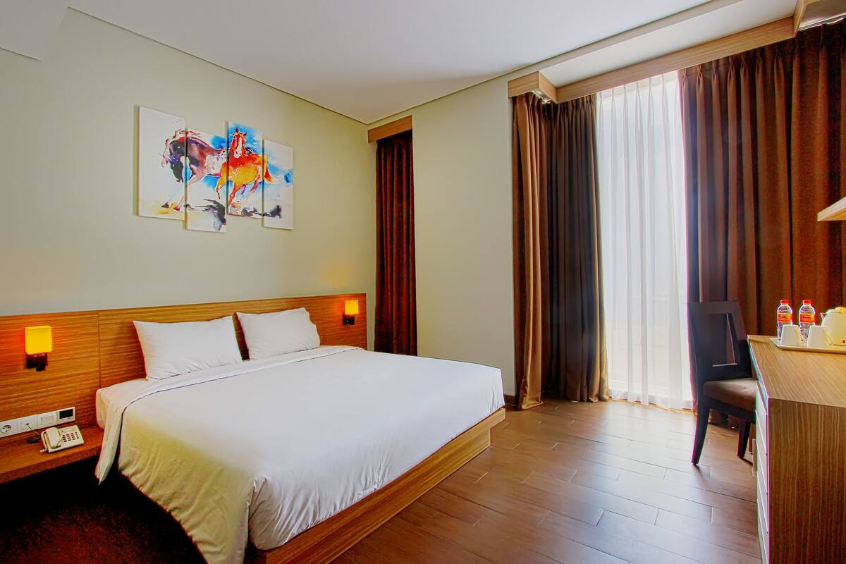 A hotel room with a large bed, a desk with a chair, a painting on the wall, and a window with curtains.