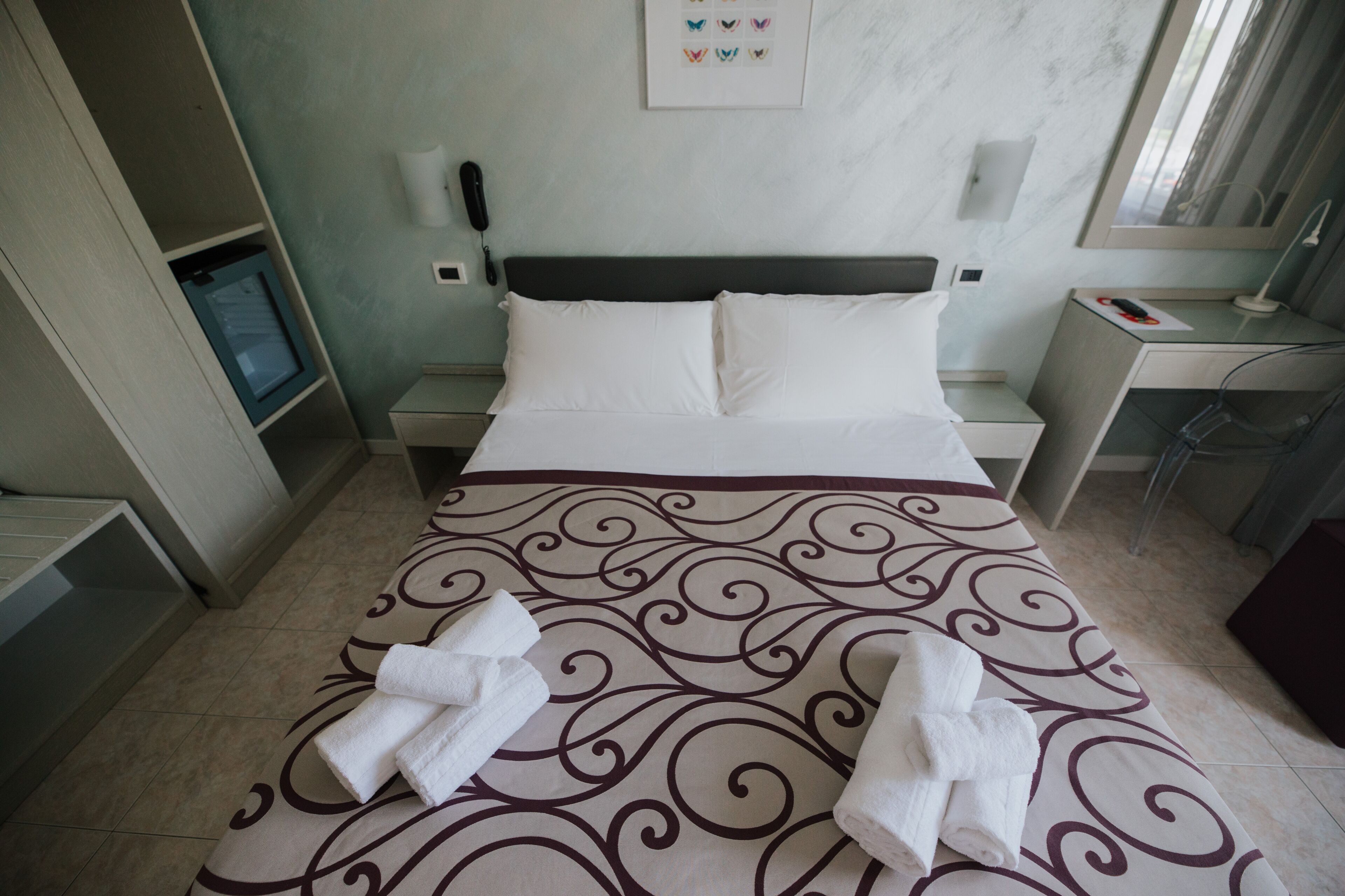 A neatly made bed with a patterned bedspread and two towels. There is a bedside table with a lamp, a phone, and a small shelf. The room has a light blue wall and a framed picture above the bed.