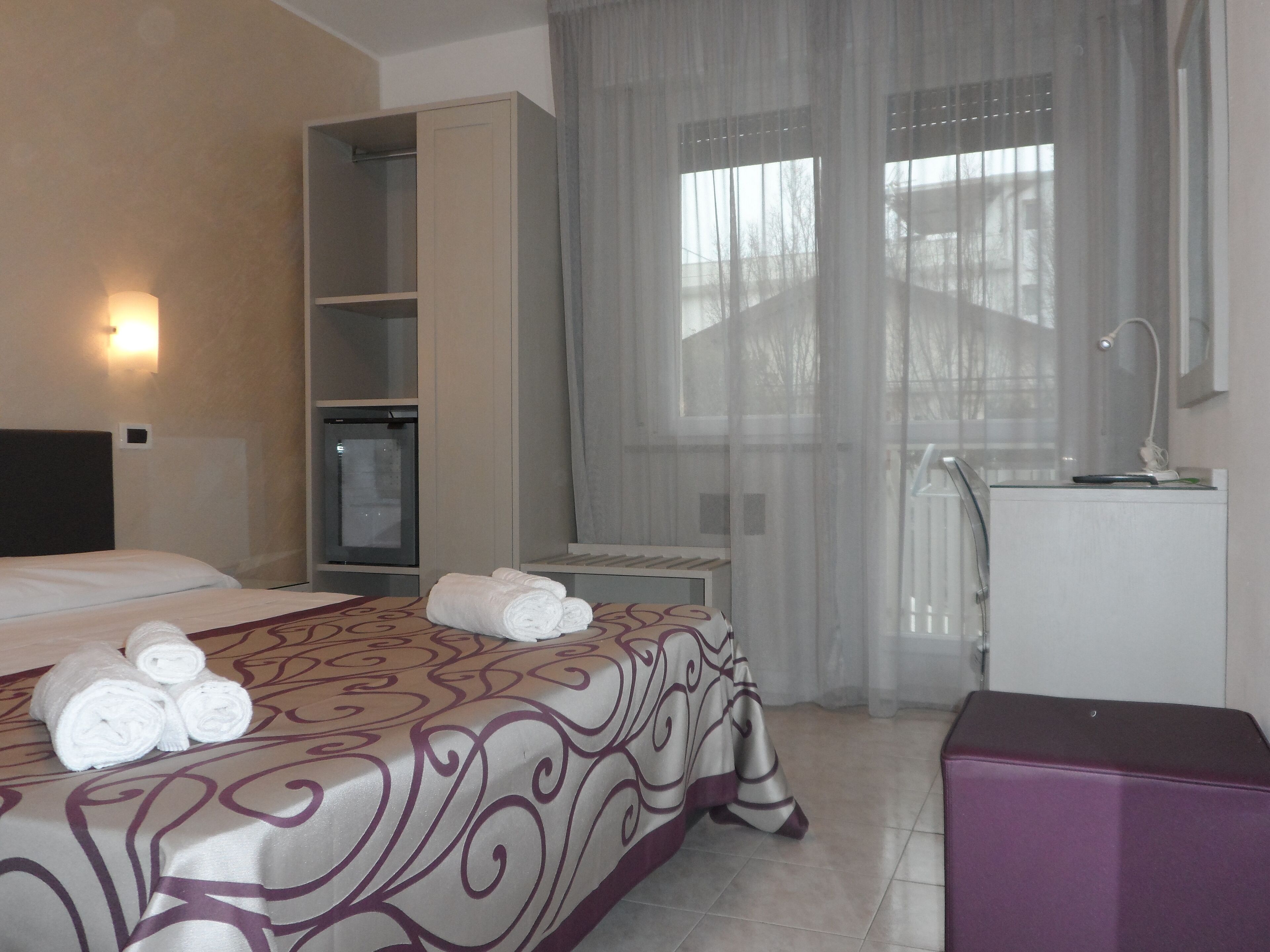 A hotel room with a bed, a desk, a wardrobe, and a window with sheer curtains.