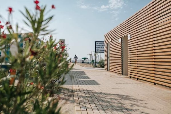 A wooden building with a sloped roof, a paved walkway, and a sign in the background. - Hotel Gloria (Lignano Sabbiadoro)