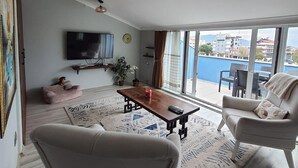 A living room with a TV, a wooden coffee table, two sofas, a large window with a view of buildings and a body of water. - Golden Moon Hotel (Fethiye)