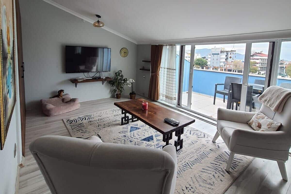 A living room with a TV, a wooden coffee table, two sofas, a large window with a view of buildings and a body of water.